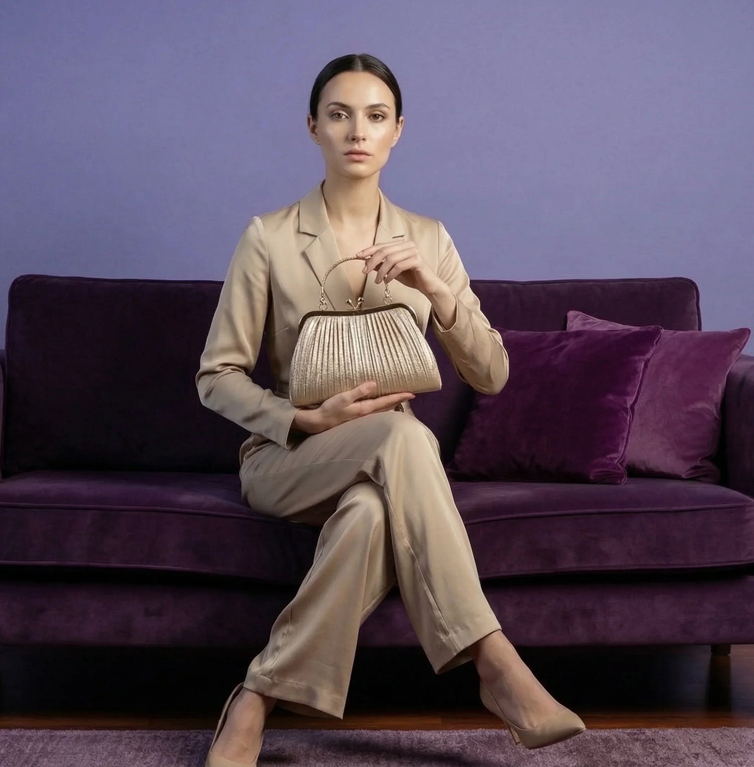 Donna in completo beige con borsa dorata a pieghe, seduta su divano viola.